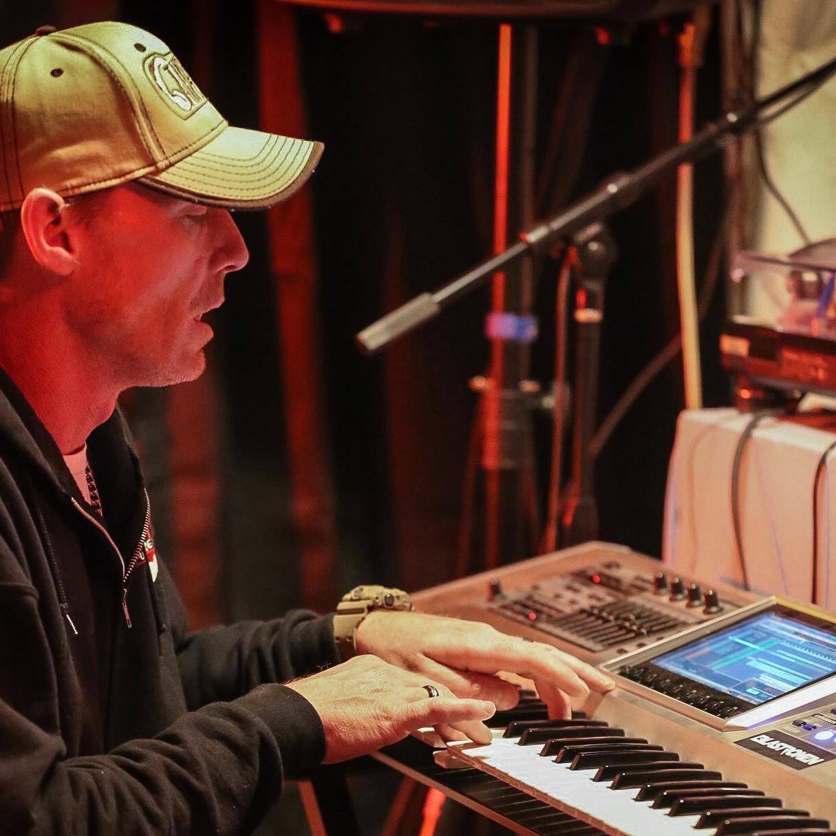 DJ Natural Nate® in side profile under warm red stage lighting playing the Roland Fantom keyboard